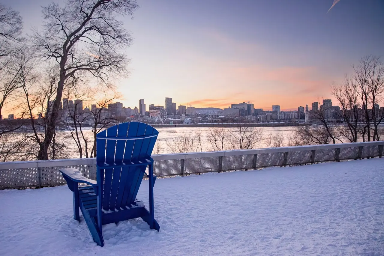 Chaise Adirondack bleue dans la neige face au fleuve et à une ville au crépuscule